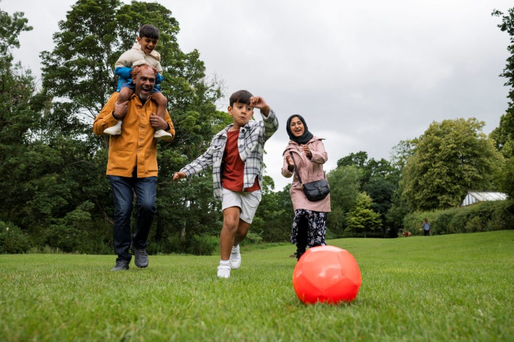 A boy runs to kick a large bright orange ball on the grass as a man with a boy on his shoulders and a woman laugh in the background.