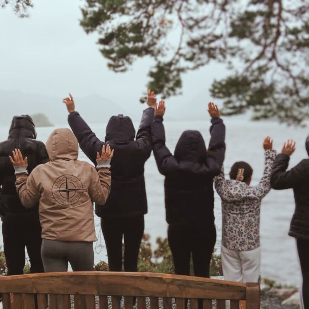 A group of young carers in warm coats raise their hands in celebration, looking out over the water.