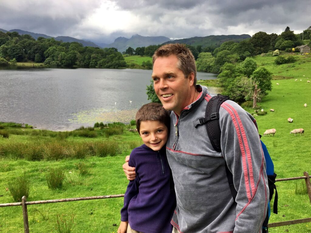A dad with his son outdoors with sheep and a lake in the background and his arm around his son, smiling