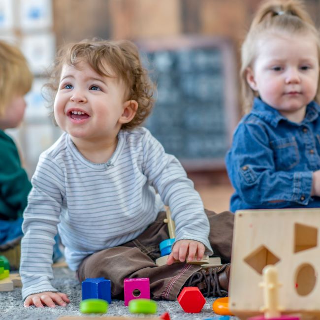 nursery-aged children sit playing with wooden blocks