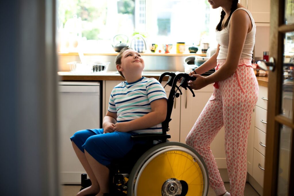 A teenage girl in pink pyjamas help her brother who is in a wheelchair with bright yellow wheels, in the kitchen.