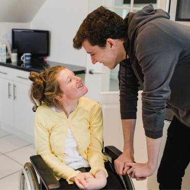 A girl in a wheelchair looks up at a teenage boy, both are smiling at each other