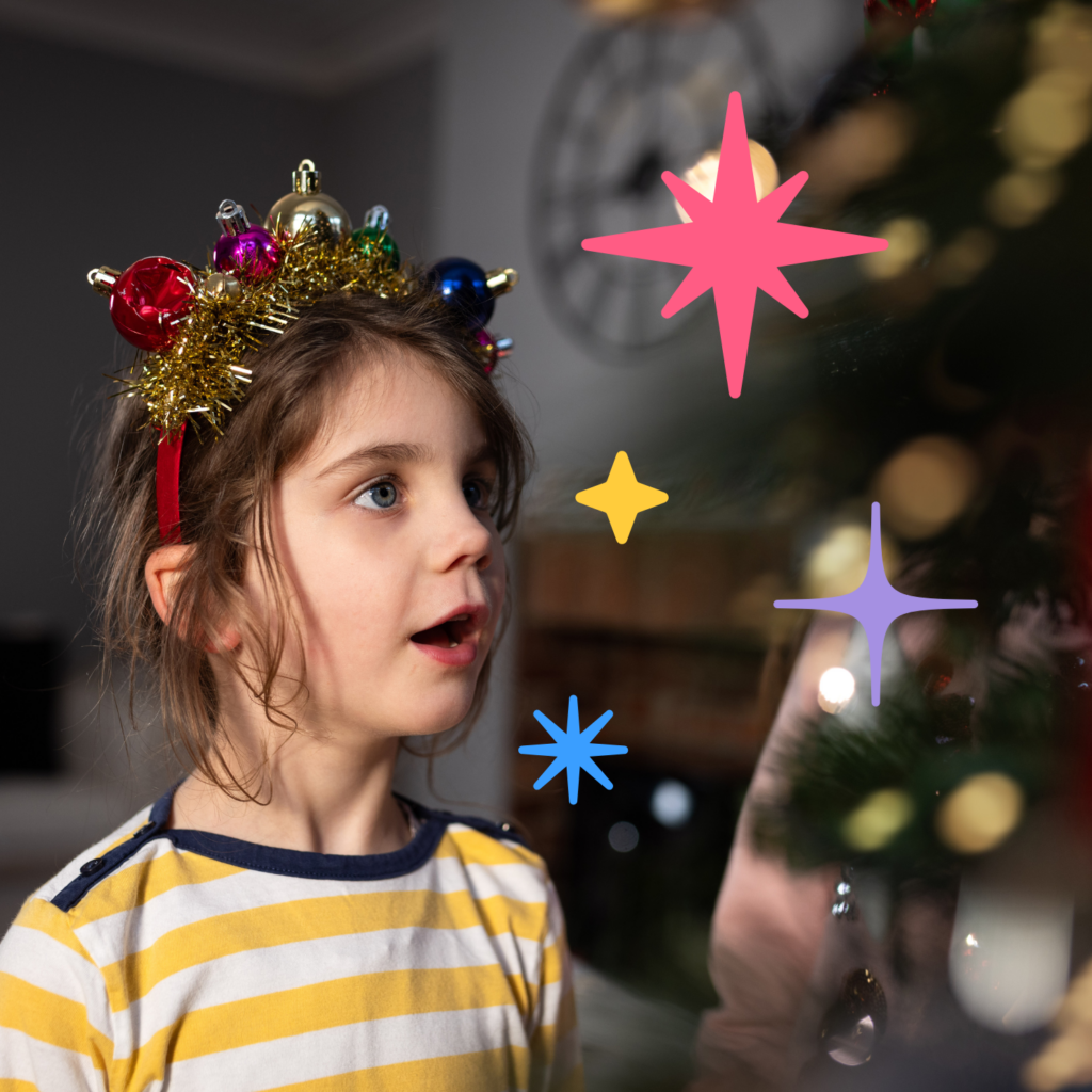 A little girl in a sparkly Christmas hairband covered in baubles looks open mouthed at a Christmas tree.
